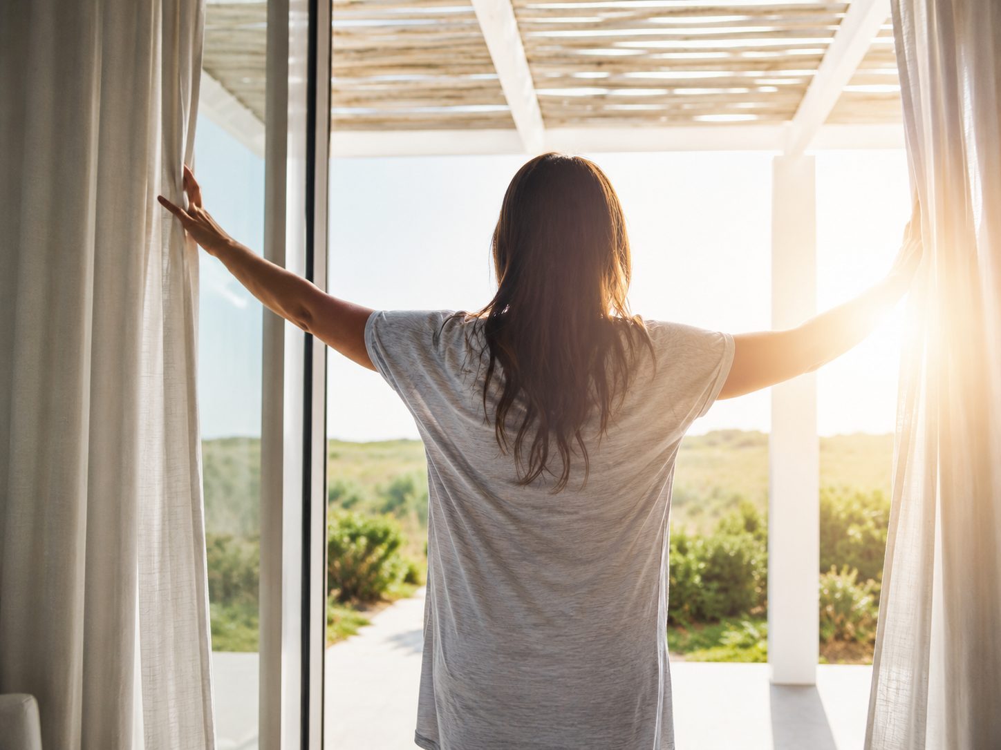 Eine Frau zieht am Morgen die Vorhänge auf, draußen eine helle Terrasse im Sonnenlicht