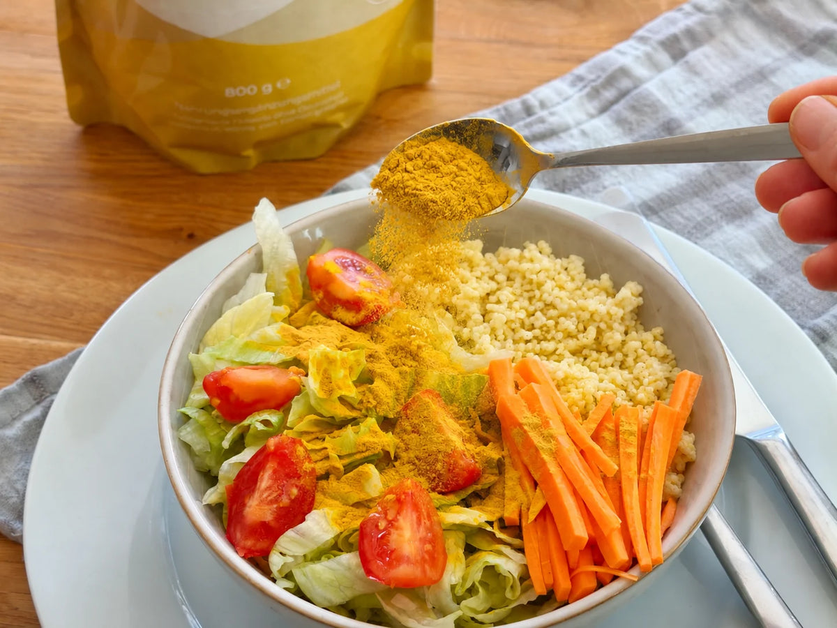 Löffel streut Umami-Gold Chlorella-Pulver über eine Schale mit Salat, Tomaten, Karotten und Couscous, dahinter der Beutel